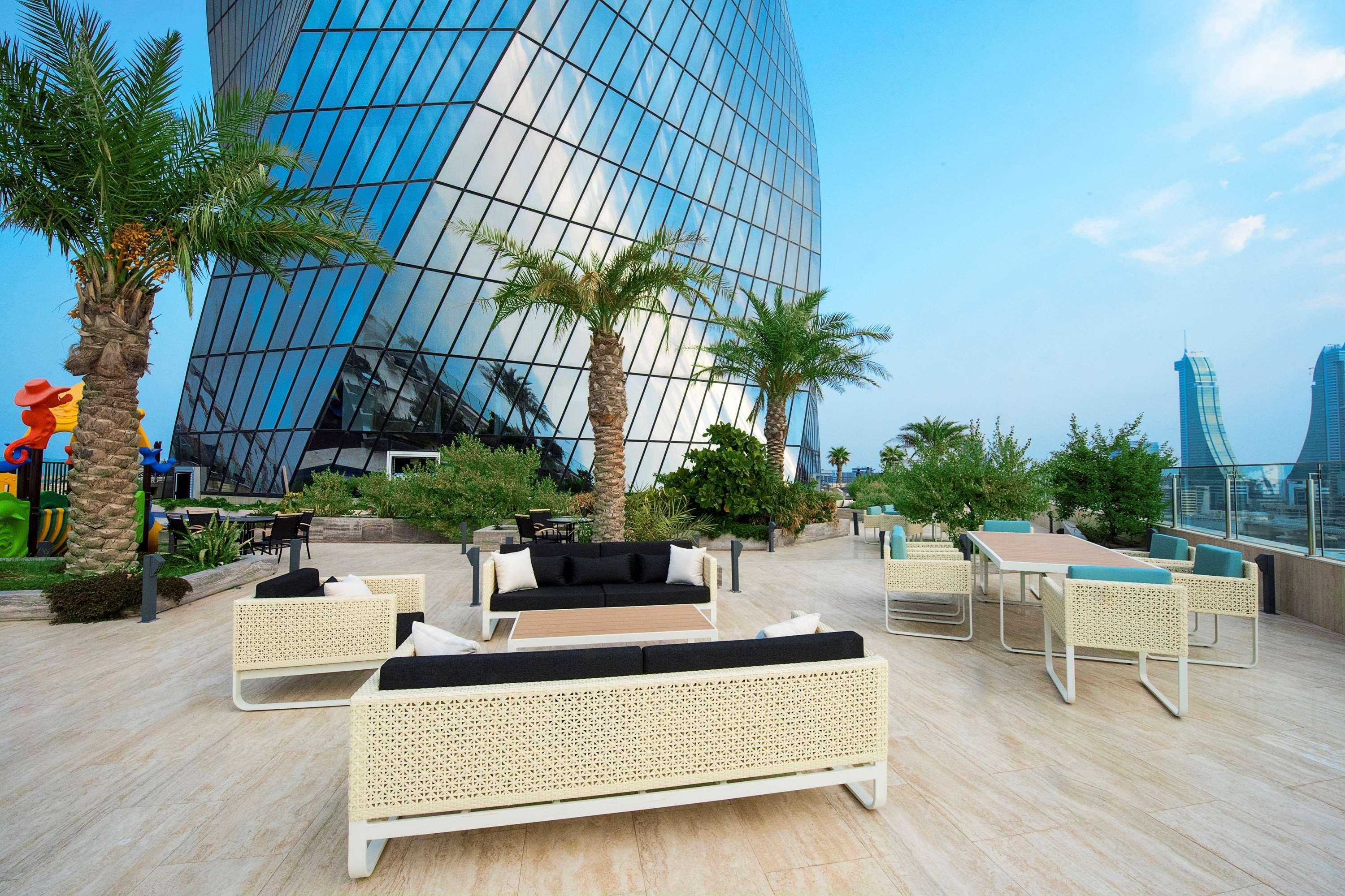 Wyndham Grand Manama Hotel Exterior photo The photo shows an outdoor seating area in front of a modern, reflective building. There are several lounge chairs and tables arranged on a light-colored tiled floor, surrounded by greenery including palm trees. The backdrop features a large glass fa