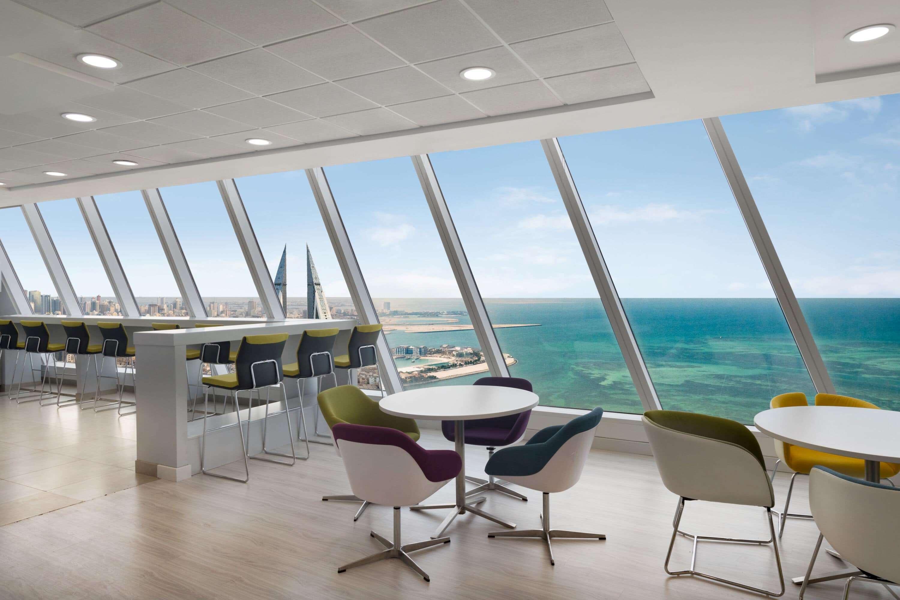 Wyndham Grand Manama Hotel Exterior photo The photo shows a modern office or lounge area with large, angled windows providing a panoramic view of the ocean and a city skyline in the distance. Inside, there are several circular tables surrounded by colorful chairs. The overall design features