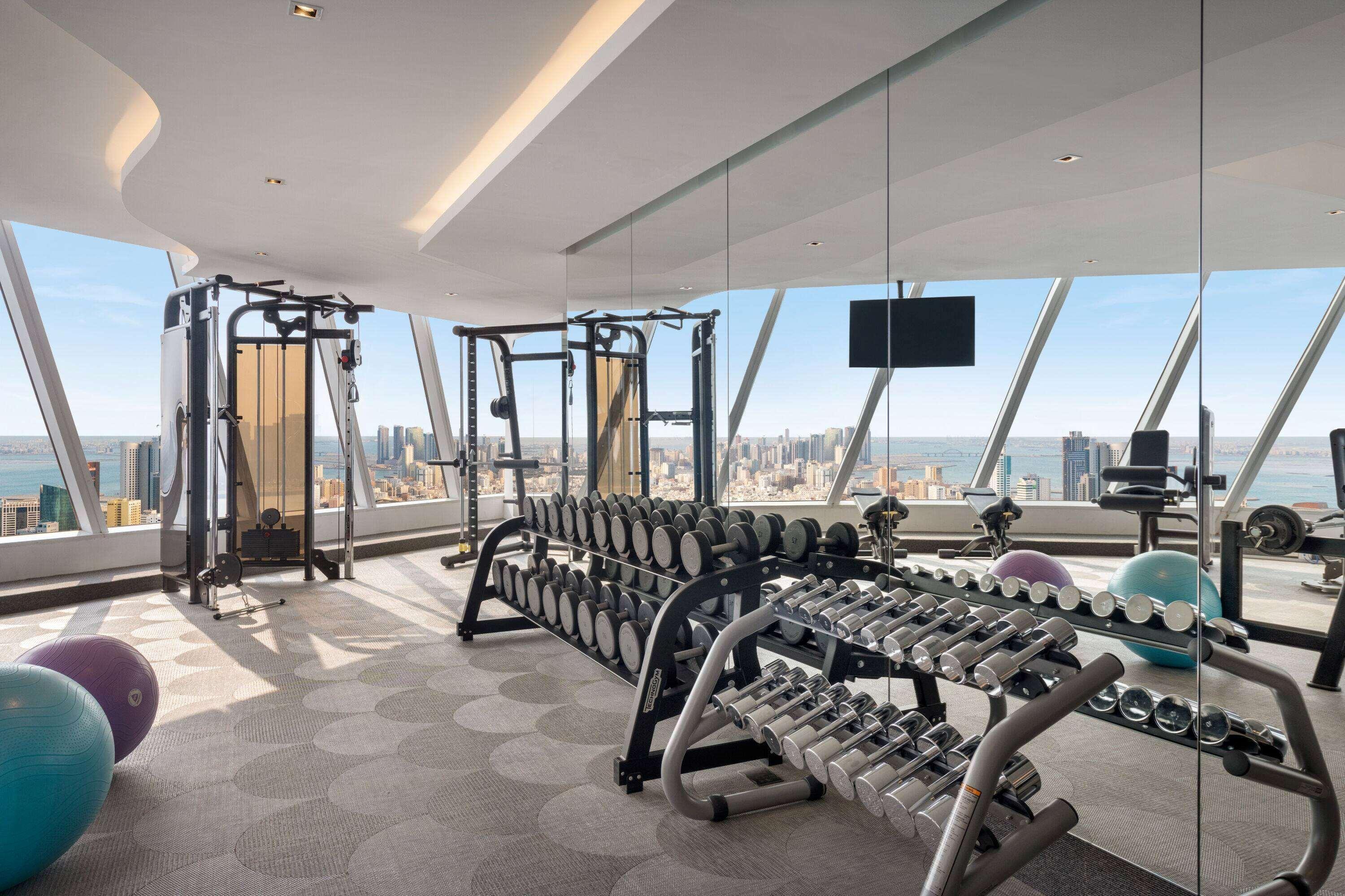 Wyndham Grand Manama Hotel Exterior photo The photo shows a modern gym with a panoramic view of a city skyline. It features various exercise equipment, including a multi-station strength training machine, a row of dumbbells on a rack, and stability balls. Large windows allow natural light to