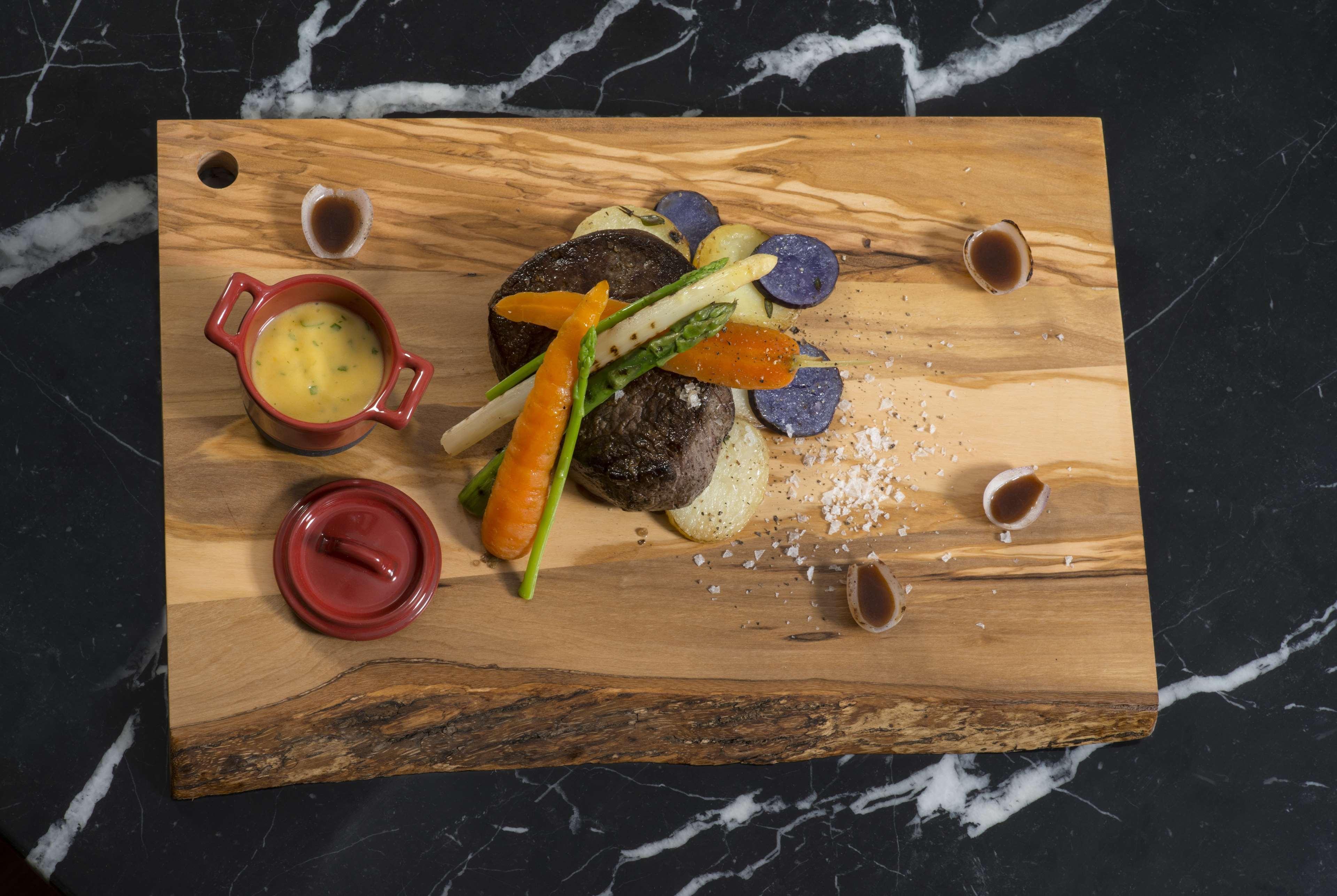 Wyndham Grand Manama Hotel Exterior photo The photo shows a beautifully plated dish on a wooden serving board. It features a portion of meat, likely beef, accompanied by several colorful vegetables, including a carrot and asparagus. There are also blue and white potato slices present. A smal