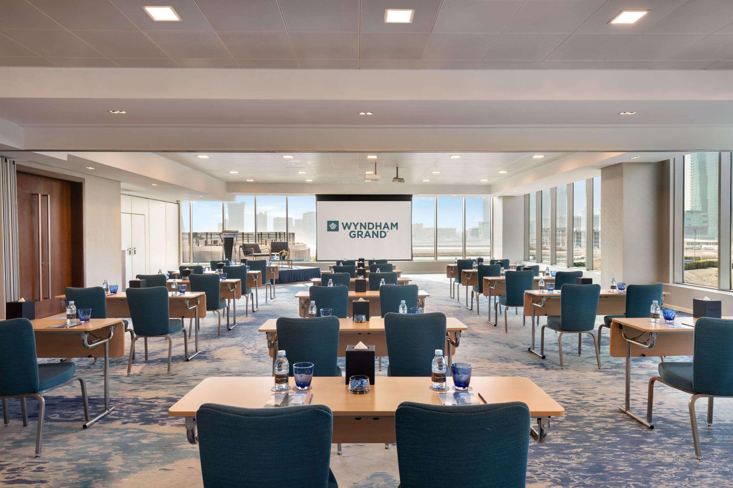 Wyndham Grand Manama Hotel Exterior photo The photo shows a modern conference room set up for a meeting. It features several rows of tables with comfortable blue chairs arranged in a classroom style. Each table has a small water bottle and note-taking supplies. At the front of the room, ther