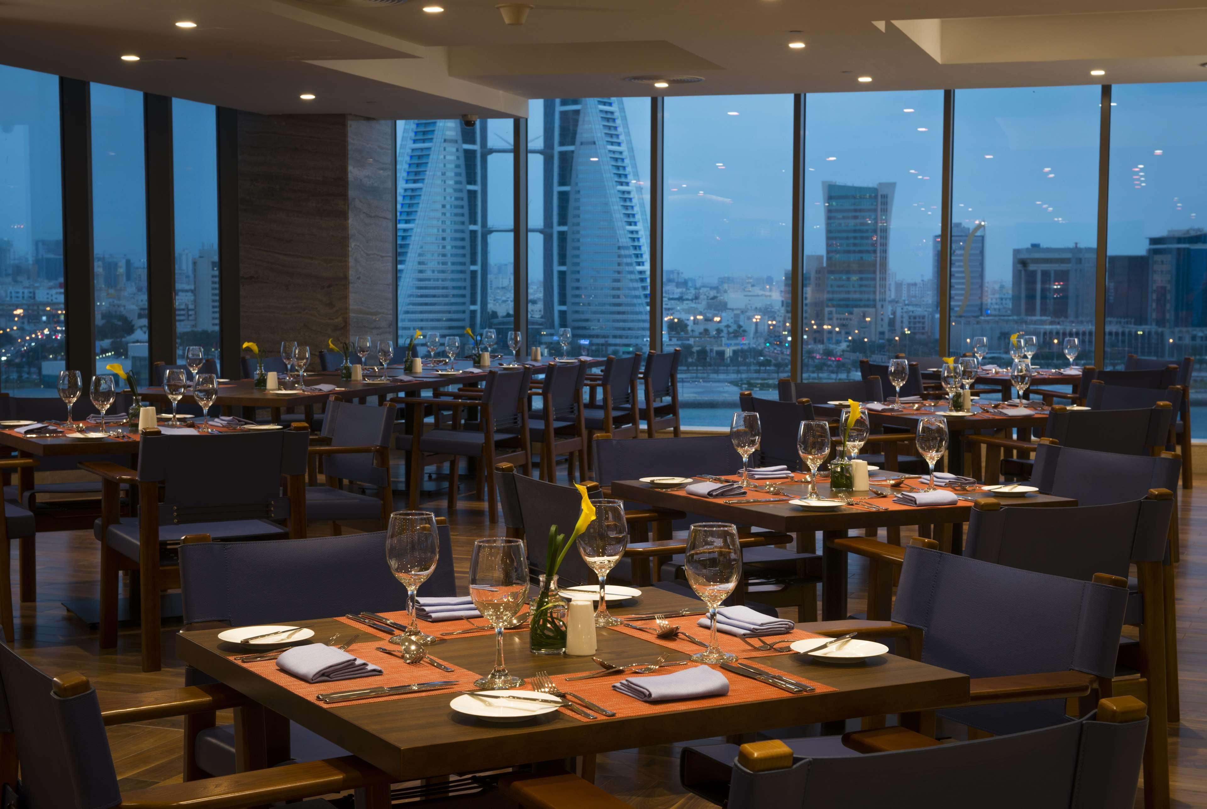 Wyndham Grand Manama Hotel Exterior photo The photo shows the interior of a restaurant, featuring multiple tables set for dining. Each table has glasses and napkins arranged neatly, and some have small floral centerpieces. Large windows offer a view of the city skyline at dusk, with building