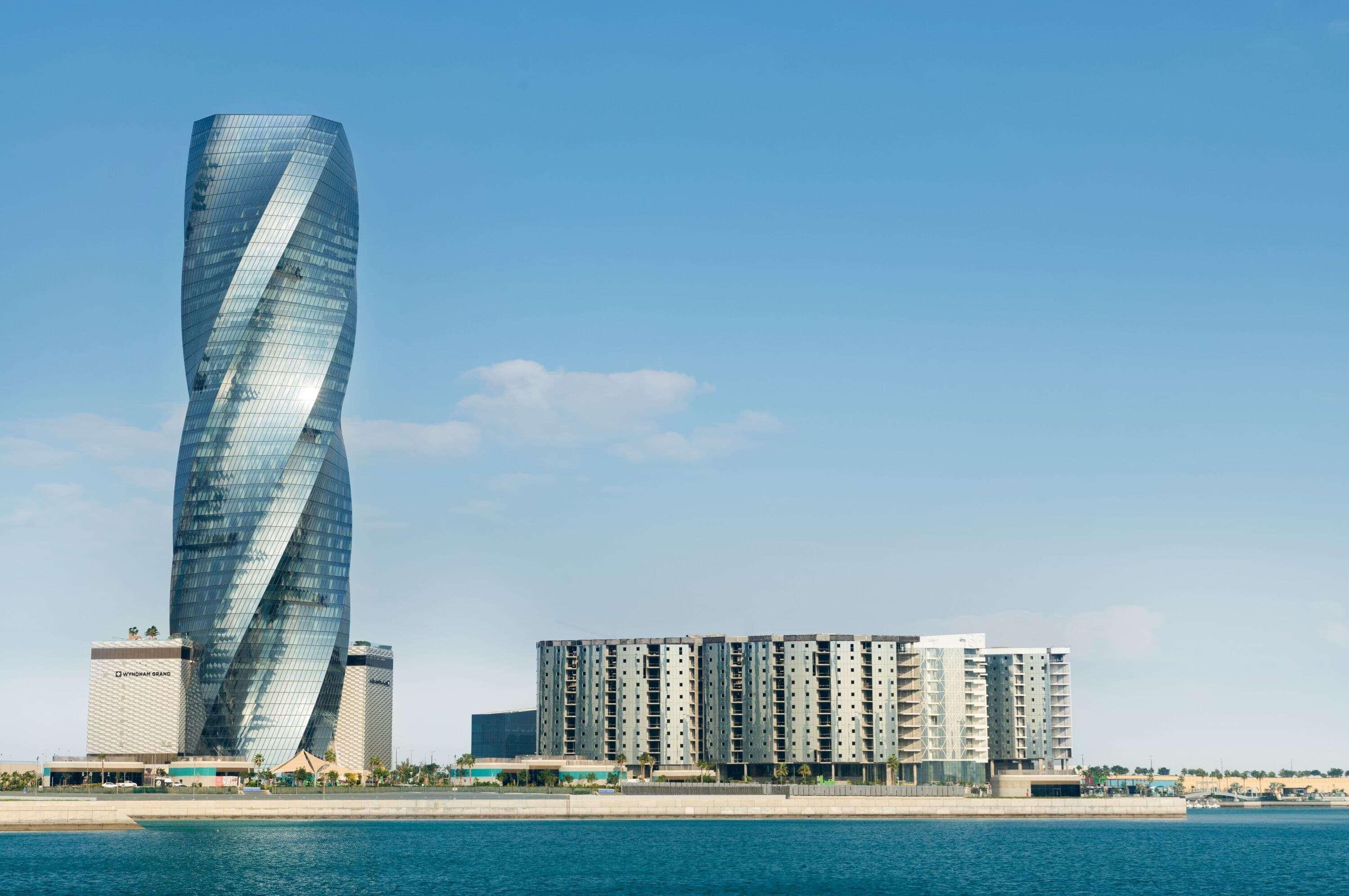 Wyndham Grand Manama Hotel Exterior photo The photo shows a modern architectural scene featuring a tall, twisting skyscraper on the left, alongside a low-rise building that appears to have multiple units. The skyscraper has a sleek, glass facade that reflects the sky, giving it a dynamic and