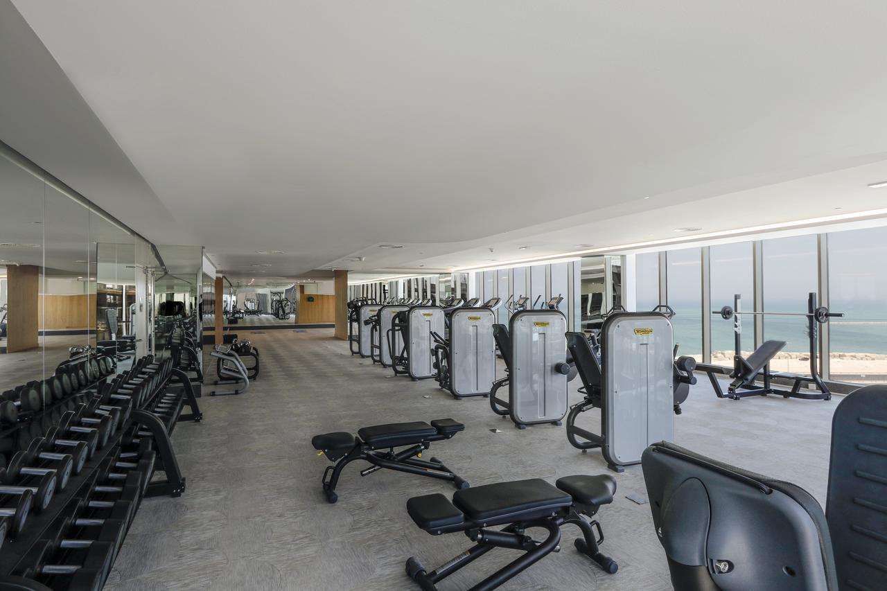 Wyndham Grand Manama Hotel Exterior photo The image shows a modern gym or fitness center. The space features various exercise equipment, including weight machines and benches. There are rows of cardio machines, possibly treadmills or ellipticals, lined up along one side. Large windows allow 